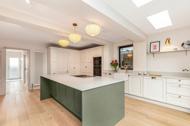 Large traditional l-shaped open plan kitchen in Other with a belfast sink, beaded cabinets, beige cabinets, quartz worktops, grey splashback, black appliances, light hardwood flooring, an island, brown floors and grey worktops.