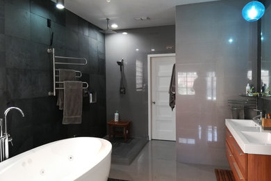 Modern master bathroom with a freestanding tub, an open shower, a two-piece toilet, stone tile, grey walls, porcelain floors, a wall-mount sink, grey floor, an open shower, shaker cabinets, medium wood cabinets, gray tile and solid surface benchtops.
