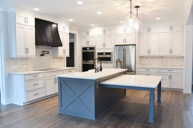 Example of a large farmhouse galley dark wood floor and brown floor eat-in kitchen design in Raleigh with a farmhouse sink, shaker cabinets, white cabinets, quartz countertops, white backsplash, glass tile backsplash, stainless steel appliances, an island and white countertops