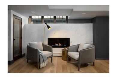 ミルウォーキーにある高級な中くらいなトランジショナルスタイルのおしゃれな地下室 (半地下 (窓あり) 、ホームバー、グレーの壁、クッションフロア、横長型暖炉、漆喰の暖炉まわり、茶色い床) の写真