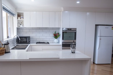 Design ideas for a mid-sized modern u-shaped eat-in kitchen in Melbourne with a double-bowl sink, flat-panel cabinets, white cabinets, white splashback, subway tile splashback, stainless steel appliances, light hardwood floors, a peninsula, granite benchtops and white benchtop.