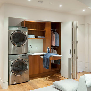 Diseño de armario lavadero lineal, contemporáneo, con fregadero bajoencimera, armarios con paneles lisos, puertas de armario de madera oscura, paredes blancas, suelo de madera en tonos medios, lavadora y secadora apiladas y encimeras blancas