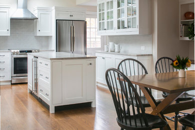 Example of a mid-sized cottage l-shaped dark wood floor eat-in kitchen design in New York with a farmhouse sink, glass-front cabinets, white cabinets, granite countertops, white backsplash, ceramic backsplash, stainless steel appliances and an island