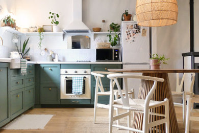 This is an example of a mid-sized beach style l-shaped open plan kitchen in Nantes with a farmhouse sink, green cabinets, laminate benchtops, beige splashback, ceramic splashback, white appliances, light hardwood floors and white benchtop.