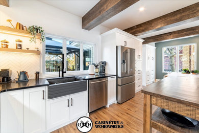 Inspiration for a mid-sized country l-shaped light wood floor, brown floor and exposed beam eat-in kitchen remodel in Toronto with a farmhouse sink, shaker cabinets, white cabinets, quartzite countertops, white backsplash, subway tile backsplash, stainless steel appliances, an island and black countertops