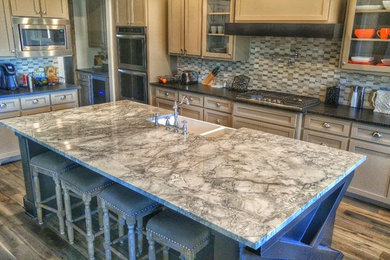 Example of a mid-sized tuscan l-shaped medium tone wood floor open concept kitchen design in Austin with a farmhouse sink, shaker cabinets, beige cabinets, granite countertops, gray backsplash, stone tile backsplash, stainless steel appliances and an island