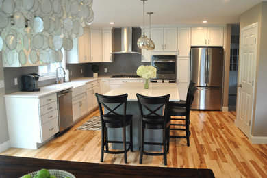 Example of a large transitional l-shaped light wood floor eat-in kitchen design in Burlington with a farmhouse sink, shaker cabinets, white cabinets, quartz countertops, gray backsplash, ceramic backsplash, stainless steel appliances and an island