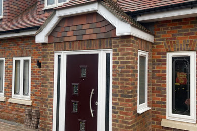 This is an example of a small rustic porch in Surrey with white walls, porcelain flooring, a single front door, a brown front door, grey floors, a vaulted ceiling, brick walls and feature lighting.