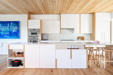 Foto på ett funkis vit kök med öppen planlösning, med en undermonterad diskho, släta luckor, vita skåp, bänkskiva i kvarts, vitt stänkskydd, glaspanel som stänkskydd, integrerade vitvaror, ljust trägolv och en köksö