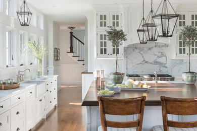 This is an example of a beach style kitchen in Boston with a farmhouse sink, white cabinets, white splashback, stone slab splashback, stainless steel appliances, medium hardwood floors, with island, white benchtop and recessed-panel cabinets.