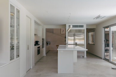 Large modern galley open plan kitchen in Central Coast with an integrated sink, flat-panel cabinets, beige cabinets, quartz benchtops, beige splashback, glass sheet splashback, black appliances, porcelain floors, with island, beige floor and white benchtop.