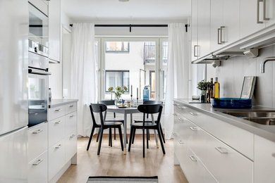 This is an example of a scandinavian galley eat-in kitchen in Malmo with a double-bowl sink, flat-panel cabinets, grey splashback, stainless steel appliances, medium hardwood floors, no island, brown floor and grey benchtop.