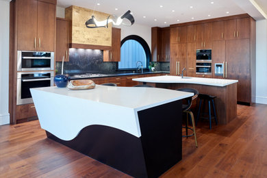Large contemporary kitchen in Vancouver with an undermount sink, flat-panel cabinets, medium wood cabinets, quartz benchtops, black splashback, stone slab splashback, panelled appliances, medium hardwood floors, multiple islands, brown floor and white benchtop.