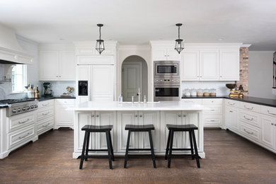 Example of a classic u-shaped dark wood floor kitchen design in Dallas with raised-panel cabinets, white cabinets, white backsplash, subway tile backsplash, an island, a farmhouse sink and stainless steel appliances
