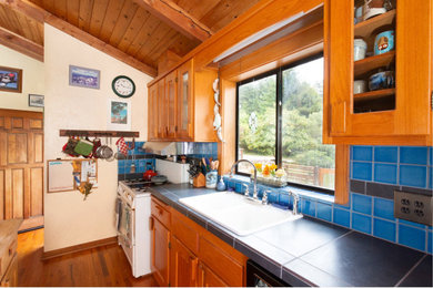 Mid-sized mountain style galley medium tone wood floor open concept kitchen photo in Other with a double-bowl sink, shaker cabinets, medium tone wood cabinets, tile countertops, blue backsplash, ceramic backsplash, an island and black countertops