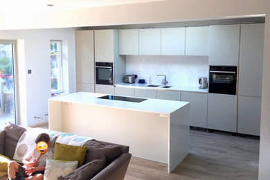 Inspiration for a medium sized modern galley open plan kitchen in Sussex with a submerged sink, grey cabinets, quartz worktops, metallic splashback, glass sheet splashback, black appliances, laminate floors, an island, brown floors and white worktops.