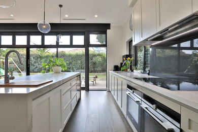 Inspiration for a mid-sized galley open plan kitchen in Sydney with an undermount sink, shaker cabinets, white cabinets, quartz benchtops, black splashback, glass sheet splashback, stainless steel appliances, medium hardwood floors, with island, brown floor and white benchtop.