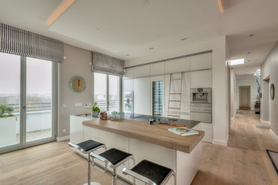 Photo of a large modern galley open plan kitchen in Berlin with a drop-in sink, flat-panel cabinets, white cabinets, stainless steel benchtops, white splashback, glass sheet splashback, stainless steel appliances, medium hardwood floors, with island, brown floor and grey benchtop.