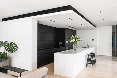Photo of a mid-sized contemporary single-wall open plan kitchen in Perth with a single-bowl sink, flat-panel cabinets, black cabinets, quartz benchtops, black splashback, stainless steel appliances, light hardwood floors, with island, brown floor and white benchtop.