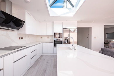 This is an example of a large modern grey and white l-shaped open plan kitchen in London with white cabinets, quartz worktops, white splashback, stainless steel appliances, an island, grey floors and white worktops.