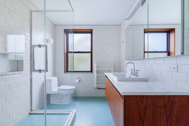 Minimalist blue tile mosaic tile floor corner shower photo in New York with flat-panel cabinets, medium tone wood cabinets, a one-piece toilet, white walls, a drop-in sink and marble countertops
