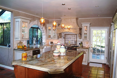 Inspiration for a mid-sized mediterranean l-shaped terra-cotta tile and red floor kitchen remodel in San Francisco with a farmhouse sink, raised-panel cabinets, distressed cabinets, granite countertops, beige backsplash, stone tile backsplash and stainless steel appliances