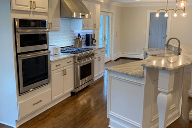 Large transitional l-shaped medium tone wood floor and brown floor open concept kitchen photo in Other with a farmhouse sink, raised-panel cabinets, white cabinets, granite countertops, white backsplash, ceramic backsplash, stainless steel appliances, an island and gray countertops