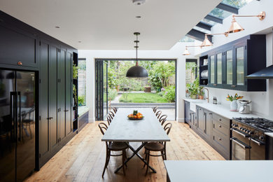 Large eclectic l-shaped kitchen/diner in London with a belfast sink, shaker cabinets, blue cabinets, engineered stone countertops, white splashback, engineered quartz splashback, black appliances, light hardwood flooring, no island, grey worktops, exposed beams and feature lighting.