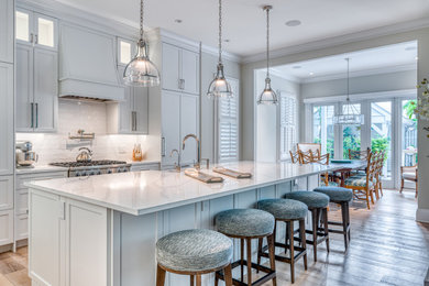 Example of a beach style medium tone wood floor and beige floor eat-in kitchen design in Miami with a farmhouse sink, shaker cabinets, white cabinets, quartz countertops, white backsplash, ceramic backsplash, stainless steel appliances, an island and white countertops