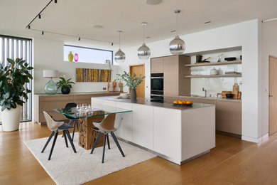 Medium sized contemporary l-shaped open plan kitchen in Sussex with a double-bowl sink, flat-panel cabinets, light wood cabinets, stainless steel worktops, white splashback, glass sheet splashback, black appliances, medium hardwood flooring and an island.