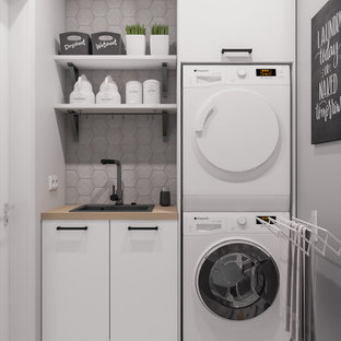 Modelo de cuarto de lavado de galera, escandinavo, pequeño, con fregadero encastrado, puertas de armario blancas, encimera de madera, suelo de baldosas de cerámica, lavadora y secadora apiladas, suelo gris, paredes grises, armarios abiertos y encimeras beige