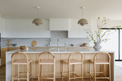 Example of a huge trendy l-shaped light wood floor and beige floor eat-in kitchen design in San Diego with an undermount sink, flat-panel cabinets, white cabinets, solid surface countertops, white backsplash, stone slab backsplash, stainless steel appliances, an island and white countertops