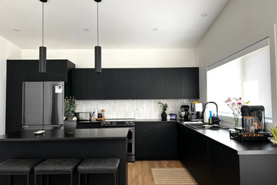 Inspiration for a mid-sized modern l-shaped medium tone wood floor and vaulted ceiling kitchen remodel in Other with flat-panel cabinets, black cabinets, white backsplash, ceramic backsplash, black appliances, an island and black countertops