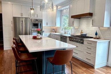 Photo of an expansive traditional l-shaped kitchen in New York with a submerged sink, shaker cabinets, white cabinets, engineered stone countertops, white splashback, ceramic splashback, stainless steel appliances, medium hardwood flooring, an island, brown floors and white worktops.