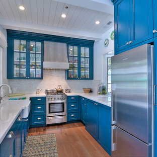 На фото: маленькие п-образные, отдельные кухни в морском стиле с раковиной в стиле кантри, синими фасадами, белым фартуком, техникой из нержавеющей стали, паркетным полом среднего тона, стеклянными фасадами и фартуком из удлиненной плитки