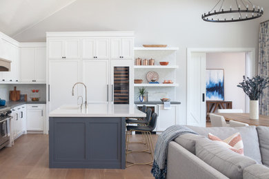 Example of a large beach style l-shaped light wood floor open concept kitchen design in Orange County with a farmhouse sink, shaker cabinets, white cabinets, quartz countertops, white backsplash, marble backsplash, paneled appliances, an island and gray countertops