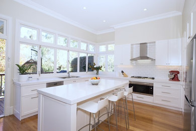 Photo of a large classic u-shaped kitchen/diner in Sydney with a double-bowl sink, shaker cabinets, white cabinets, engineered stone countertops, white splashback, metro tiled splashback, stainless steel appliances, medium hardwood flooring, an island, brown floors and white worktops.