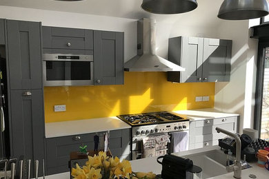 Photo of a contemporary kitchen in London with an integrated sink, shaker cabinets, grey cabinets, composite countertops, yellow splashback, glass sheet splashback and stainless steel appliances.