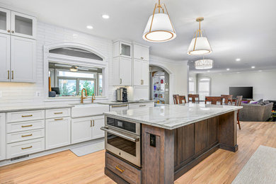 Inspiration for a transitional u-shaped kitchen in DC Metro with a farmhouse sink, shaker cabinets, white cabinets, quartzite benchtops, white splashback, ceramic splashback, stainless steel appliances, medium hardwood floors, with island, brown floor and white benchtop.