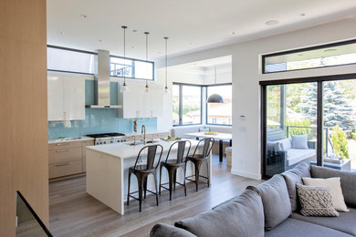Photo of a large contemporary l-shaped open plan kitchen in Vancouver with a double-bowl sink, flat-panel cabinets, light wood cabinets, quartz benchtops, blue splashback, glass sheet splashback, stainless steel appliances, medium hardwood floors, with island, grey floor and white benchtop.