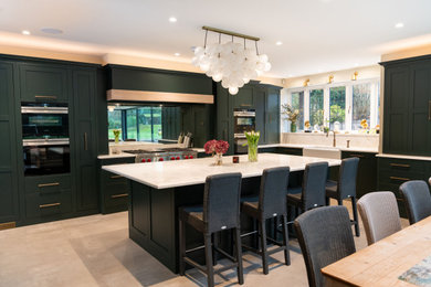 Photo of a large industrial l-shaped open plan kitchen in Essex with a farmhouse sink, flat-panel cabinets, green cabinets, quartzite benchtops, metallic splashback, mirror splashback, stainless steel appliances, limestone floors, with island, grey floor and grey benchtop.