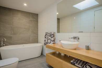 This is an example of a mid-sized contemporary master bathroom in Auckland with light wood cabinets, a freestanding tub, a one-piece toilet, gray tile, porcelain tile, white walls, porcelain floors, a vessel sink, wood benchtops and beaded inset cabinets.