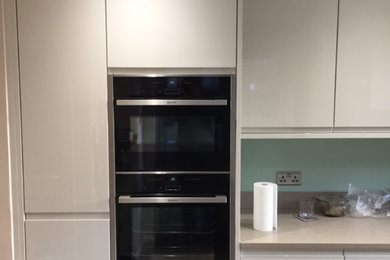 Photo of a modern enclosed kitchen in Cambridgeshire with white cabinets, quartz worktops, green splashback, glass sheet splashback, black appliances and no island.