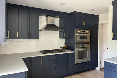 Inredning av ett klassiskt mellanstort grå grått kök, med skåp i shakerstil, blå skåp, bänkskiva i kvarts, vitt stänkskydd, stänkskydd i porslinskakel, rostfria vitvaror, ljust trägolv, en halv köksö och beiget golv