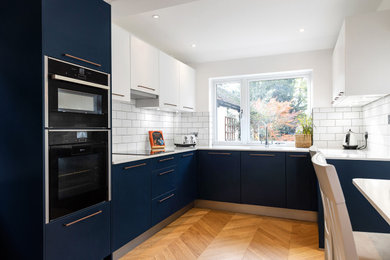 This is an example of a mid-sized contemporary u-shaped eat-in kitchen in Hertfordshire with an undermount sink, flat-panel cabinets, blue cabinets, quartzite benchtops, white splashback, ceramic splashback, black appliances, light hardwood floors, with island, brown floor and white benchtop.