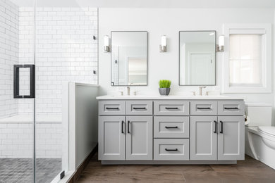 Photo of a mid-sized transitional 3/4 bathroom in DC Metro with recessed-panel cabinets, grey cabinets, an alcove shower, a two-piece toilet, white tile, subway tile, white walls, an undermount sink, brown floor, a hinged shower door, white benchtops and engineered quartz benchtops.