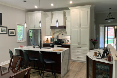 Example of a mid-sized transitional galley laminate floor and brown floor open concept kitchen design in Other with a farmhouse sink, raised-panel cabinets, white cabinets, quartz countertops, white backsplash, quartz backsplash, stainless steel appliances, an island and white countertops