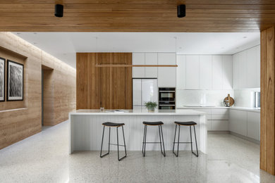 Photo of a large modern l-shaped open plan kitchen in Melbourne with a double-bowl sink, white cabinets, solid surface benchtops, white splashback, black appliances, concrete floors, with island, white floor, white benchtop and timber.