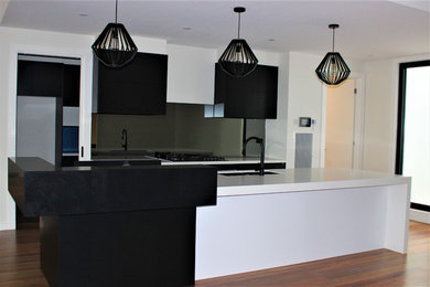 This is an example of a large contemporary single-wall open plan kitchen in Melbourne with a double-bowl sink, flat-panel cabinets, black cabinets, quartz benchtops, multi-coloured splashback, mirror splashback, black appliances, light hardwood floors, with island, beige floor and white benchtop.
