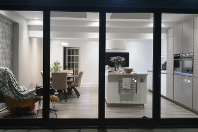 This is an example of a medium sized contemporary single-wall kitchen/diner in Other with an integrated sink, flat-panel cabinets, grey cabinets, composite countertops, metallic splashback, mirror splashback, black appliances, vinyl flooring, an island, grey floors and multicoloured worktops.